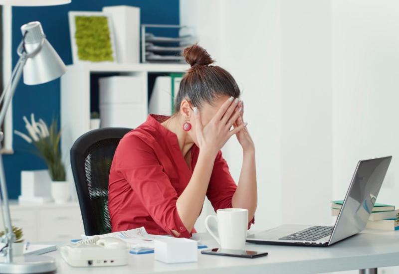 Zena u crvenoj kosulji sjedi u uredu i drzi se za glavu - 10 alarmantnih znakova burnouta na poslu i kako ih suzbiti